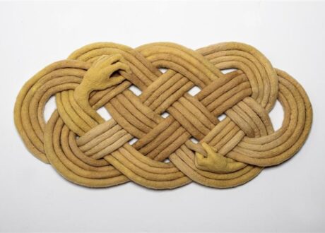 A flaxen coloured textile artwork forming a knot with two hands