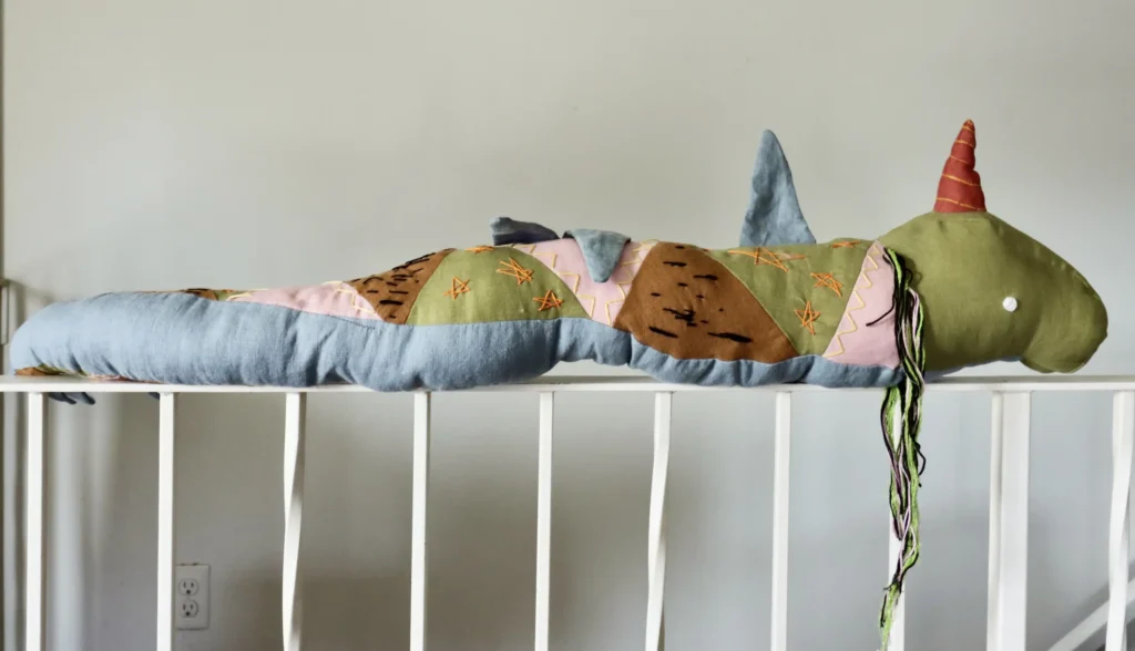 A textile artwork, Npisune’get (Provides Medicine), featuring Jipijka'm, a Mi'kmaw sea serpent, made with red, blue, black, and white fabric, embroidered with plants and symbols