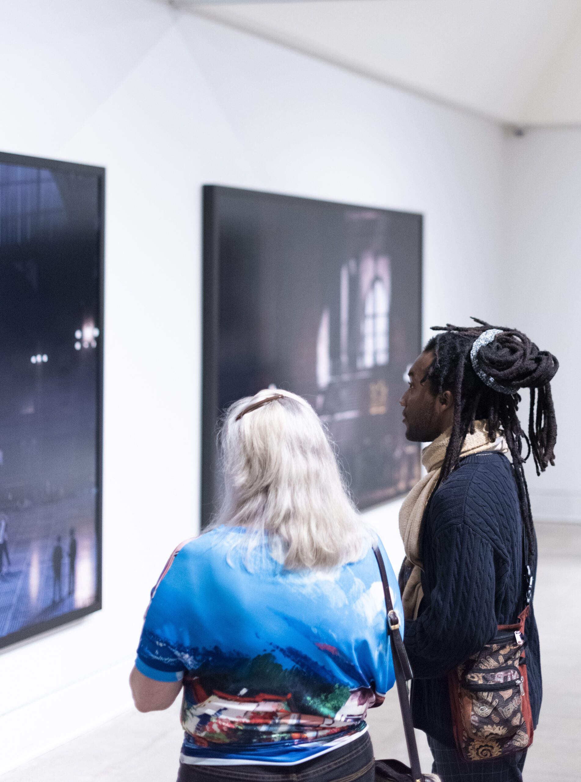 A white woman with grey hair and a Black man with dreadlocks stand in front of artwork on a white wall