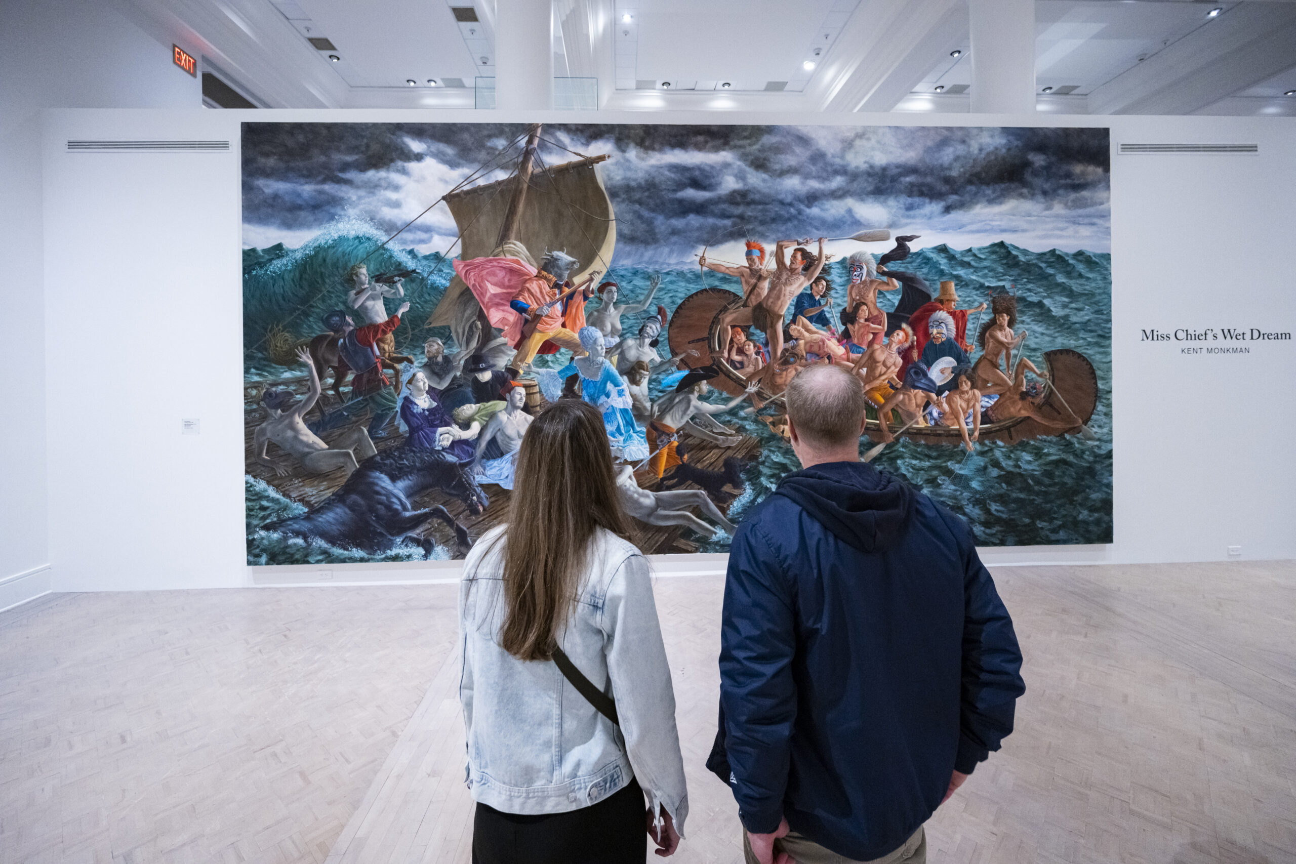 A man and a woman stand in front of a large piece of artwork by Kent Monkman