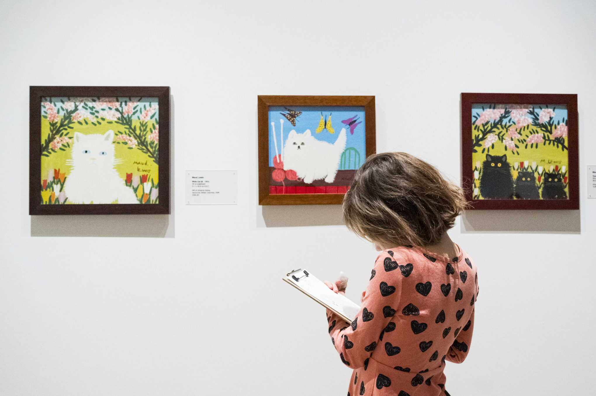 A little girl in a pink t-shirt with black hearts stands with a clipboard in front of maud lewis cat paintings