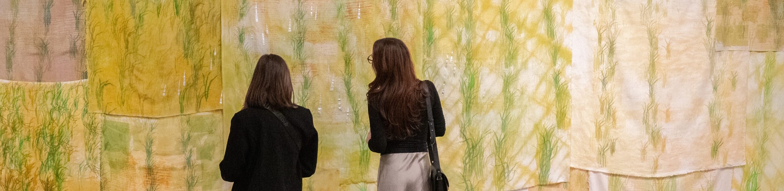 two women in dark clothing stand in front of a large piece of texile artwork
