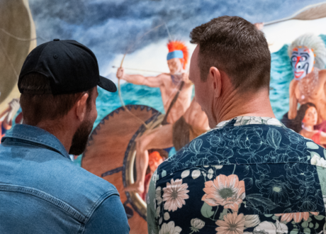 Installation view of Kent Monkman: Breaking Waves, showing two people viewed from behind looking closely at a large, vibrant painting of figures navigating stormy ocean waters in traditional boats.