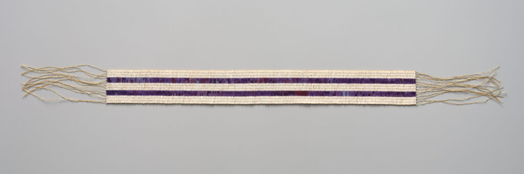 Woven Two Row Wampum belt featuring two parallel purple bead stripes set against a white beaded background, displayed horizontally with loose fringed cords at both ends.