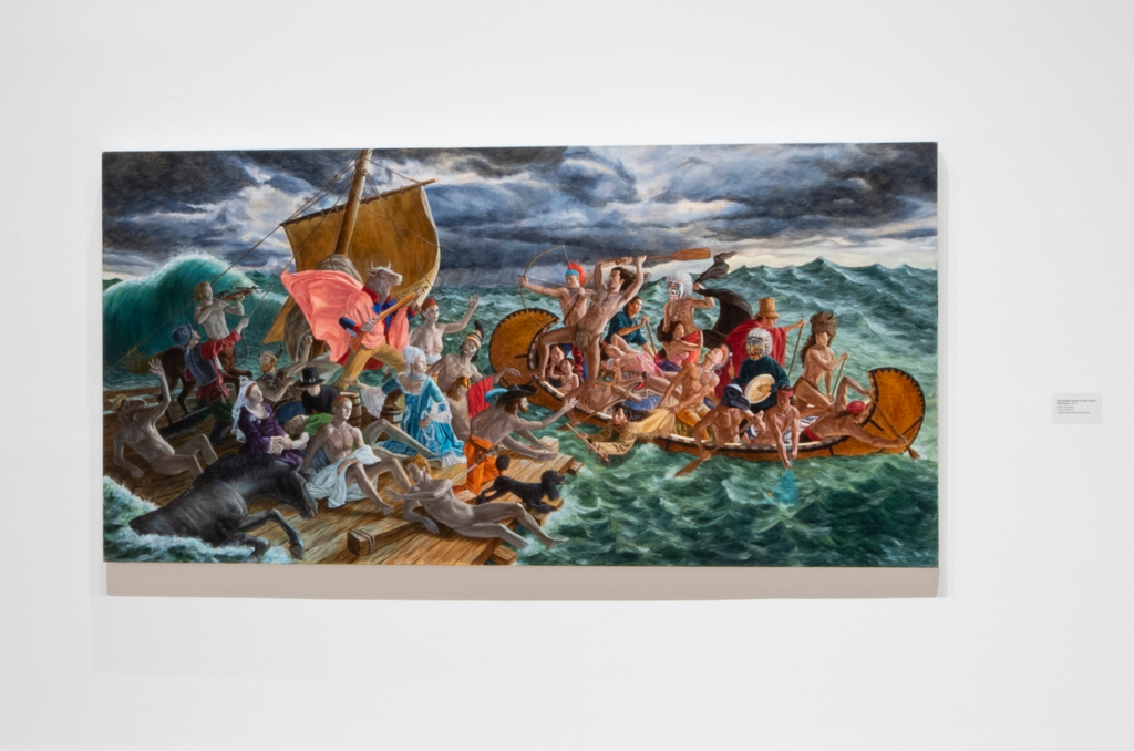 Installation view of a wide canvas painting mounted on a white gallery wall with a small exhibition wall label to its right. The painting portrays a dramatic oceanic scene under dark clouds, with Indigenous figures in a wooden canoe on the right facing European colonial figures and mythical creatures on a raft to the left.